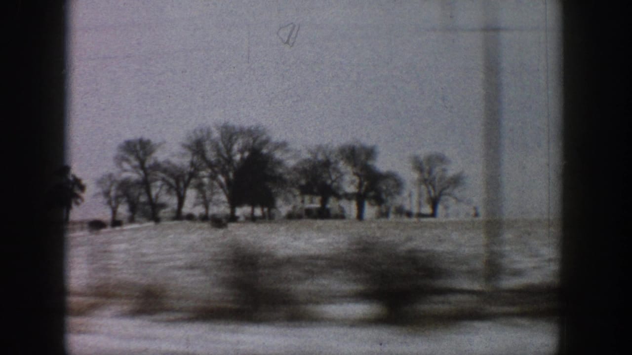 View through a car side window of snow-covered fields, bare winter trees, and distant farm outbuildings; motion blur throughout.
