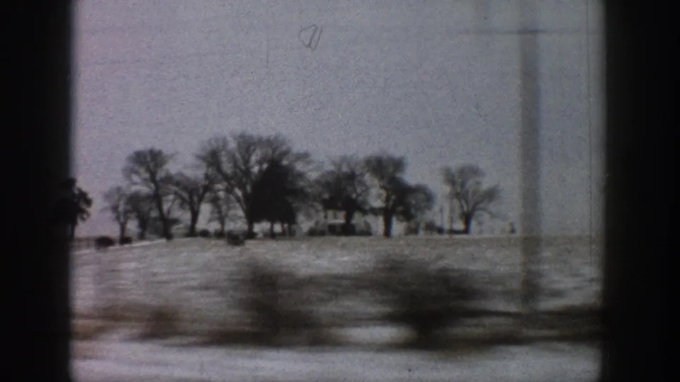 8mm home-movie side-window shot showing snow-covered fields, bare deciduous trees, and distant farmhouses; motion blur from moving vehicle; likely rural Colorado, circa 1961.