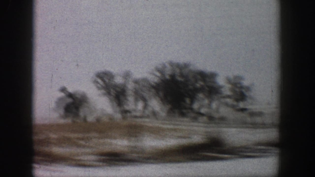 Side-window view from a moving car showing bare winter trees and a low outbuilding across snow-covered ground; motion blur in foreground.
