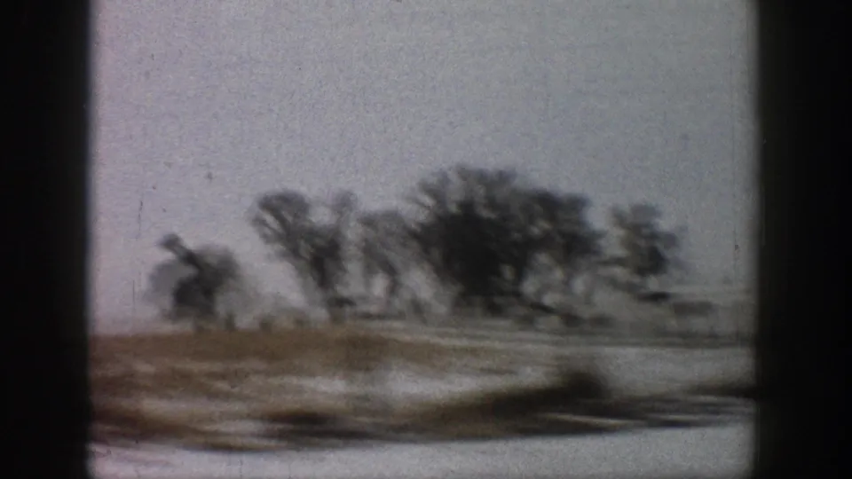 8mm side-window shot of snow-covered terrain with dense bare trees and a distant farm outbuilding; motion blur and film grain evident; rural Colorado area, circa 1961.