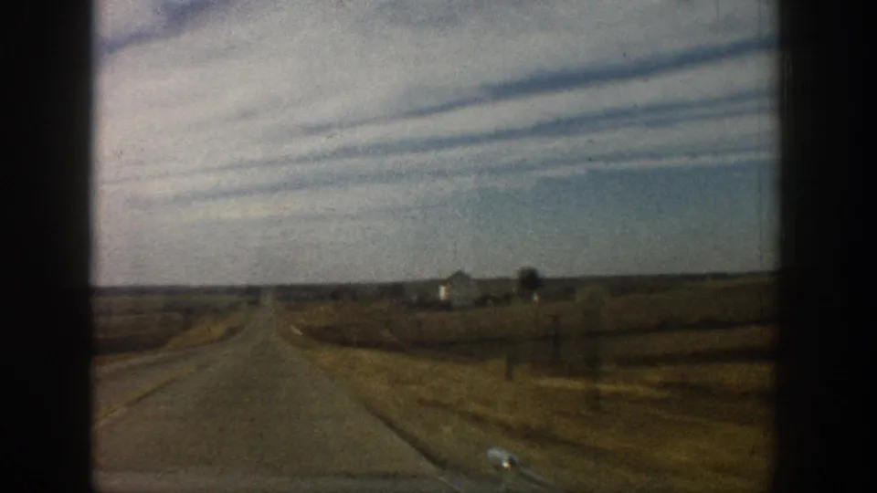 8mm forward road shot showing a straight two-lane rural road, parallel jet contrails in a blue-grey sky, utility poles, and distant farm outbuildings; circa 1961.