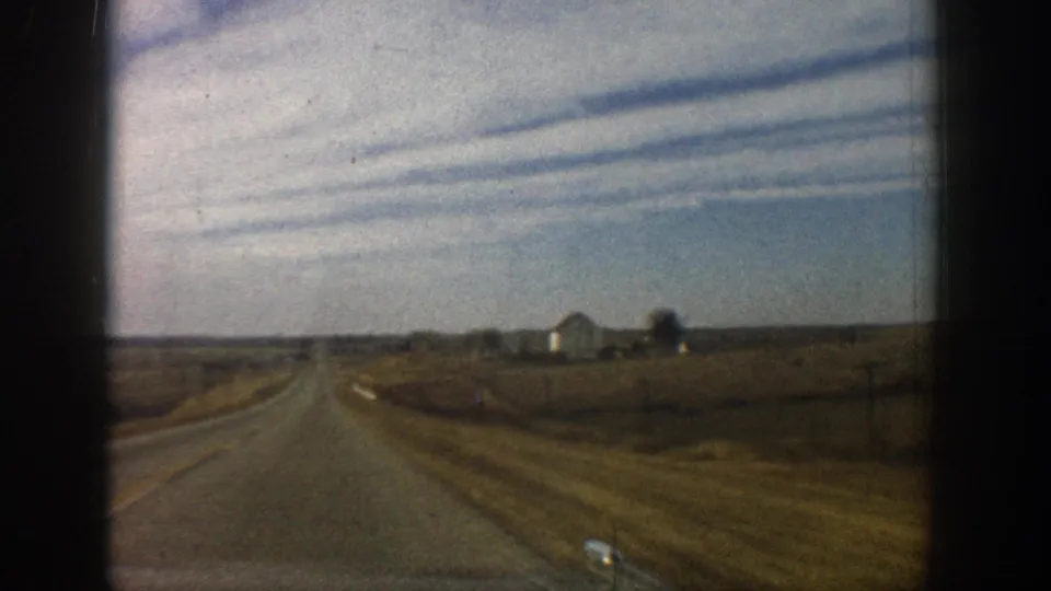 8mm windshield shot of a straight rural road with dramatic parallel jet contrails overhead, flat terrain, and a distant farm outbuilding at right; circa 1961.