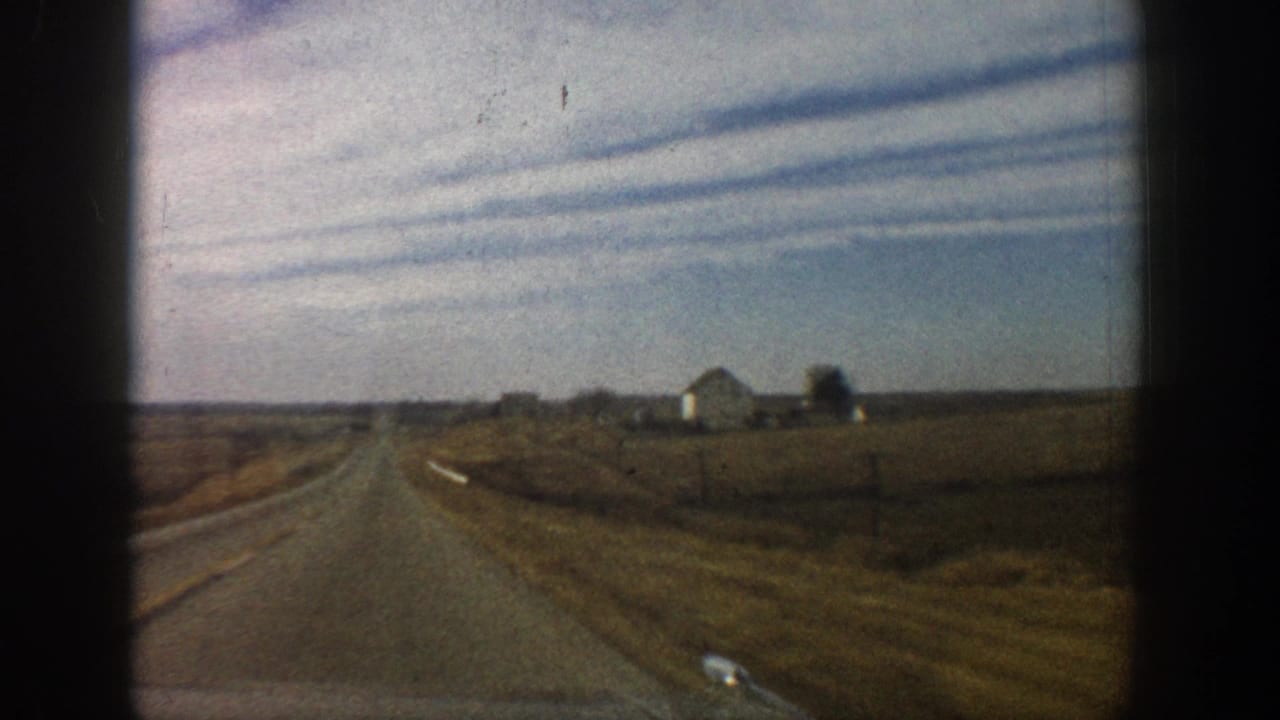 1961 8mm frame: straight two-lane road receding to the horizon with distant white farm buildings, dry golden fields either side, shot through a vehicle window.