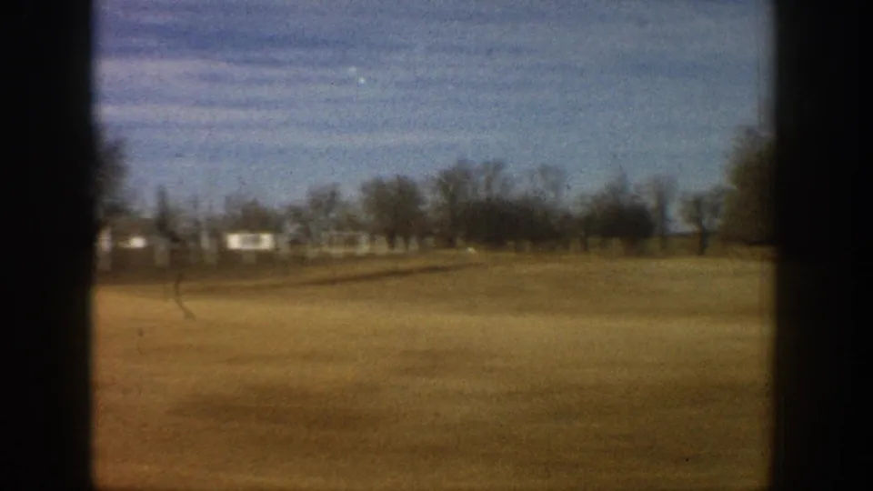 1961 8mm frame: flat open fields with bare trees and a small white farm structure at right, dry brown foreground, framed by dark window-vignette borders.