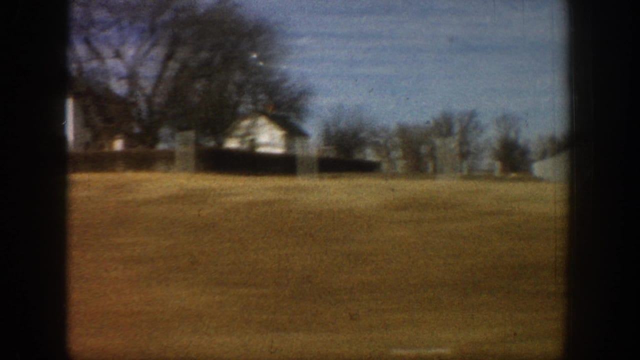 8mm home movie frame from a moving vehicle showing a white farmhouse on a gentle rise with bare trees and open brown field, rural Colorado, circa 1961.