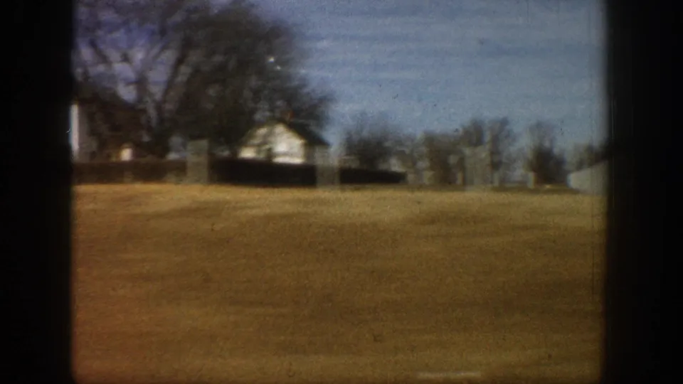 8mm home movie frame from a moving vehicle showing a white farmhouse on a gentle rise with bare trees and open brown field, rural Colorado, circa 1961.