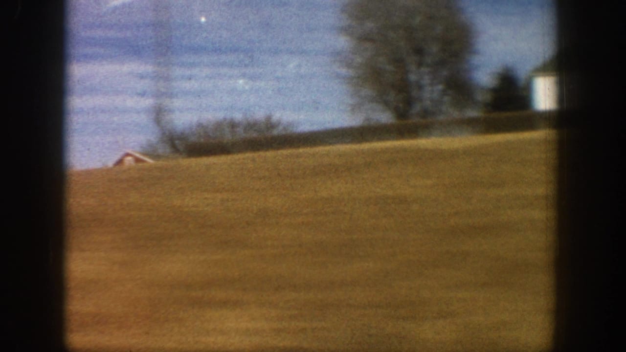8mm home movie frame from a moving vehicle showing a distant red barn on rolling golden-brown farmland with bare trees, rural Colorado, circa 1961.