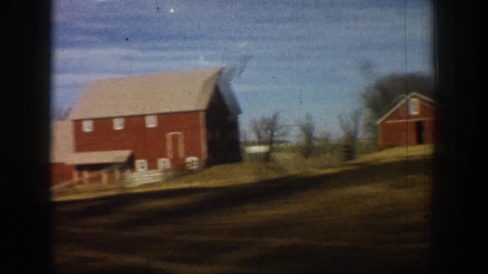 8mm home movie still showing two red barns with pale roofs on rolling brown farmland with bare trees and a fence line, rural Colorado countryside, circa 1961.