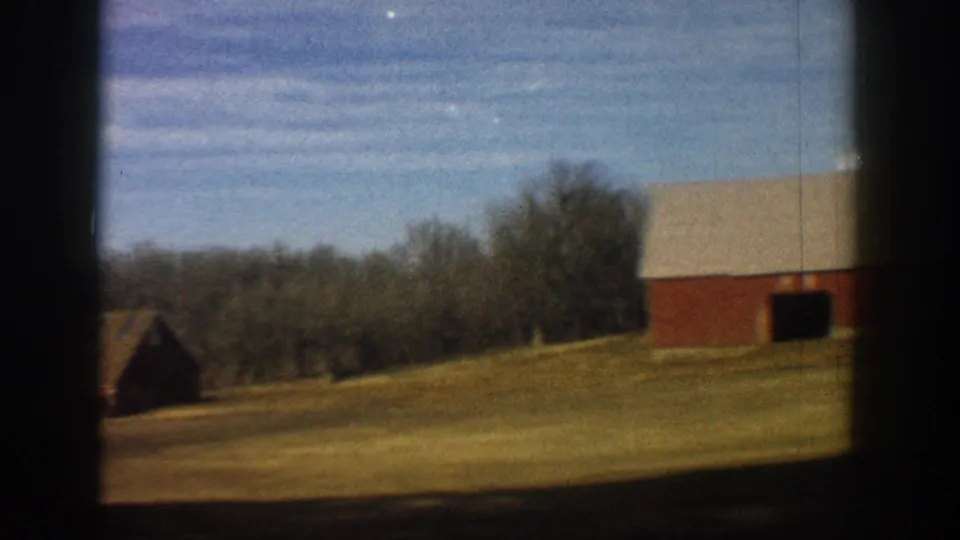 8mm home movie still showing a red barn and outbuilding on rolling golden-brown farmland with bare trees and blue sky, rural Colorado, circa 1961.