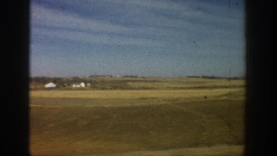 Wide open dry grassland with white farm buildings on the far horizon; 8mm road-trip footage from rural America, circa 1961.