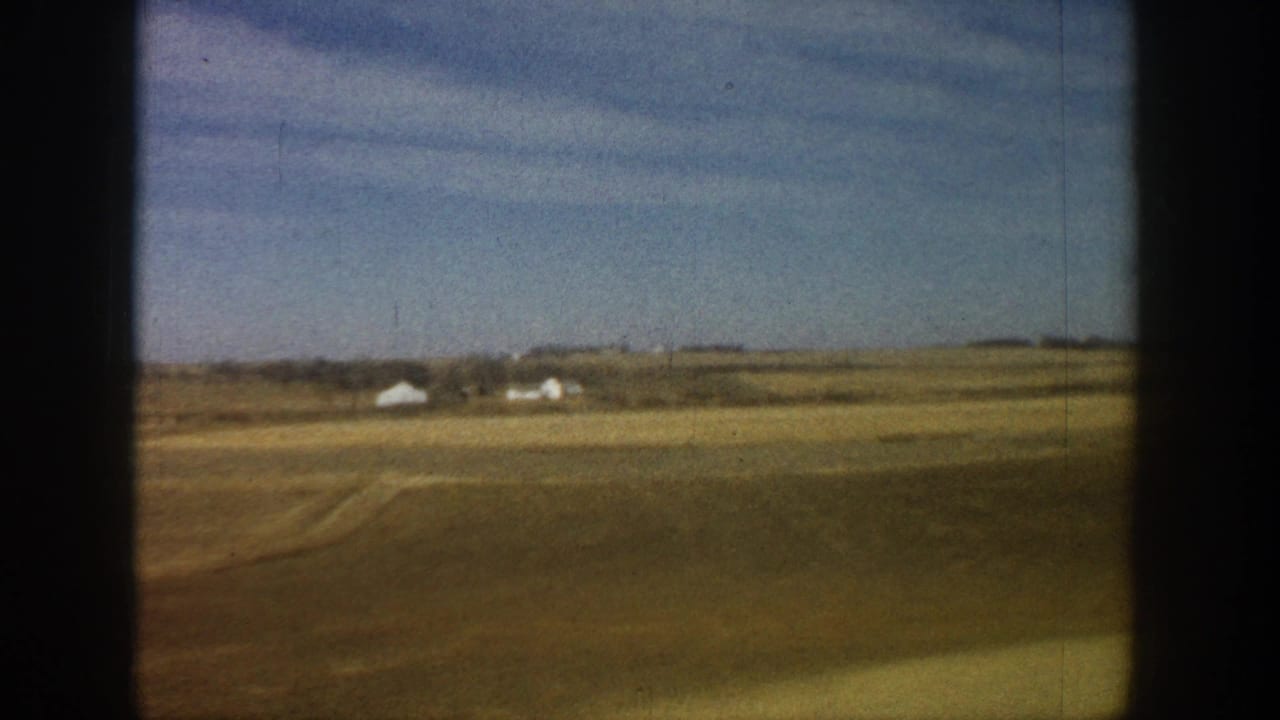 8mm home movie frame from a 1961 road trip showing vast flat plains with a white farmstead visible on the horizon.