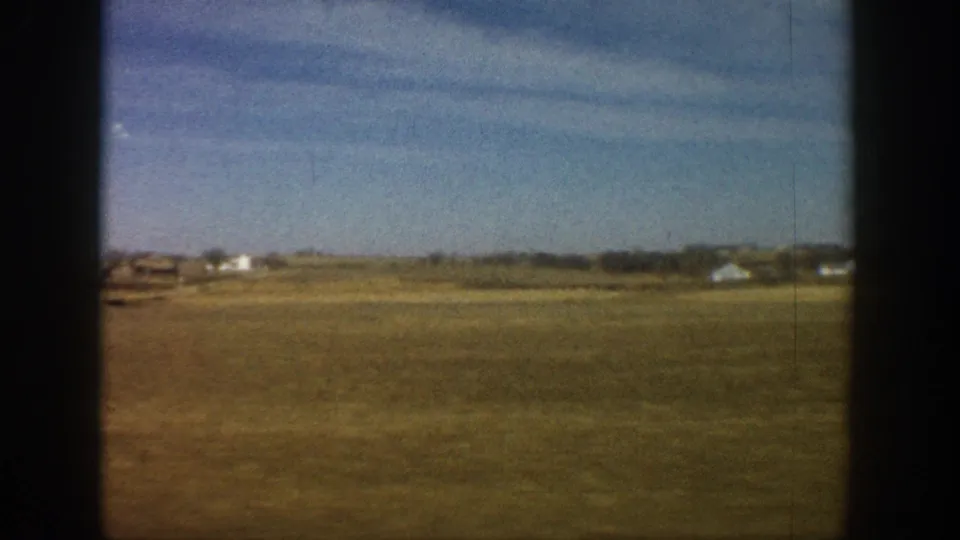 8mm home movie frame from a 1961 road trip showing flat plains, a sparse treeline, and white farm structures at the distant horizon.