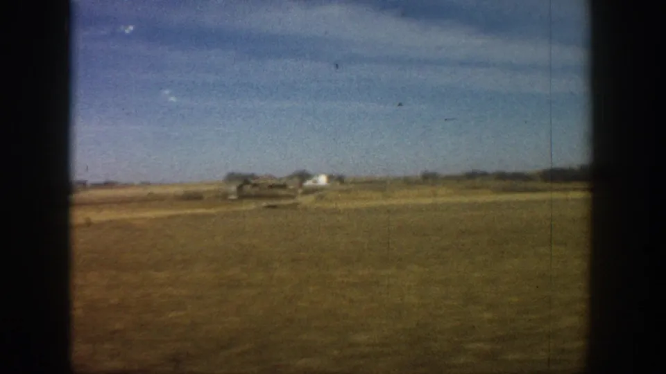 8mm travel footage from 1961 showing an expansive flat plains landscape with distant white farm buildings, viewed through a vehicle window.