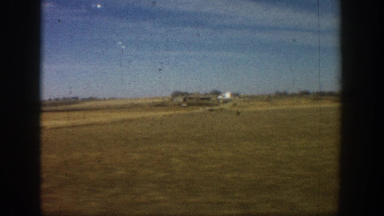 8mm home movie frame from rural Colorado, circa 1961, showing flat dry grassland and distant white farm structures under a broad blue sky.