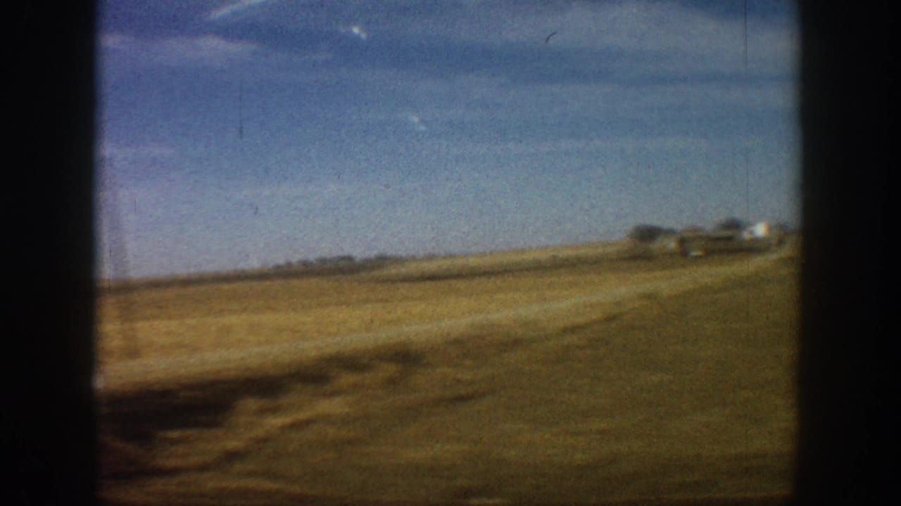 8mm home movie frame showing rural Colorado grassland from a moving vehicle, circa 1961; farm outbuildings visible on the right horizon.