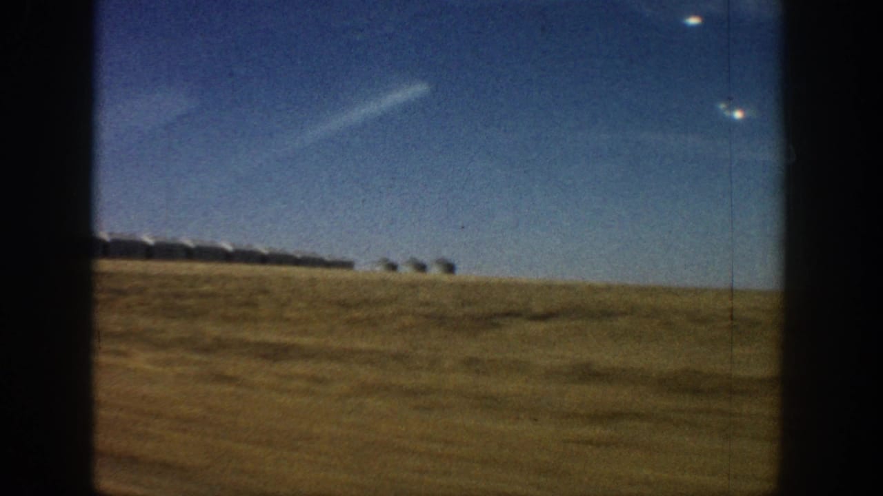8mm home movie frame circa 1961 showing a wide expanse of dry golden grassland with a distant row of white rounded-roof structures at the horizon under a deep blue Colorado sky.