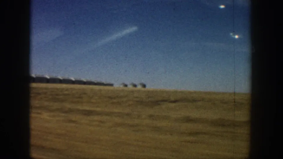 8mm home movie frame circa 1961 showing a wide expanse of dry golden grassland with a distant row of white rounded-roof structures at the horizon under a deep blue Colorado sky.