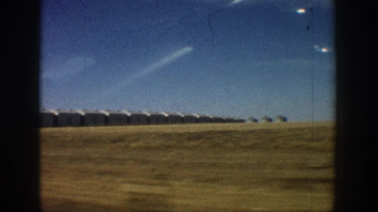 Row of white rounded-roof structures slightly closer on the horizon over flat dry grassland, with bright blue sky and film artifacts.