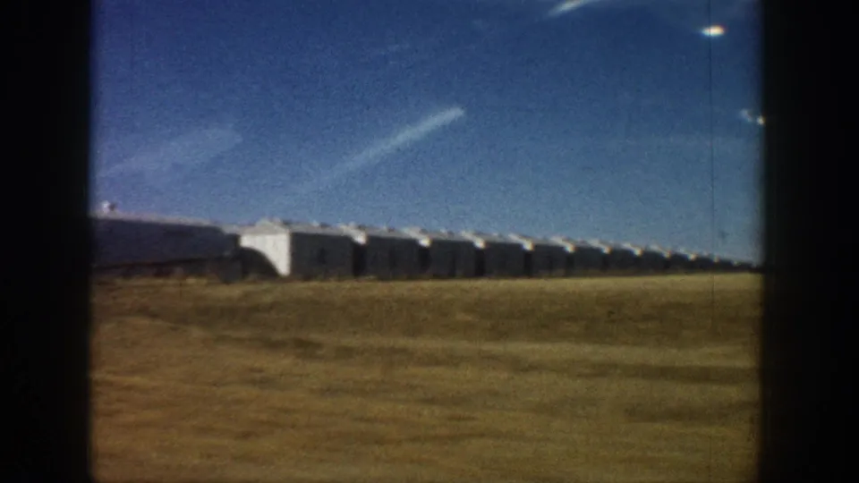 8mm home movie frame circa 1961 showing a long row of white dome or Quonset-style buildings on a Colorado plains horizon, filmed from a moving vehicle with visible motion blur.