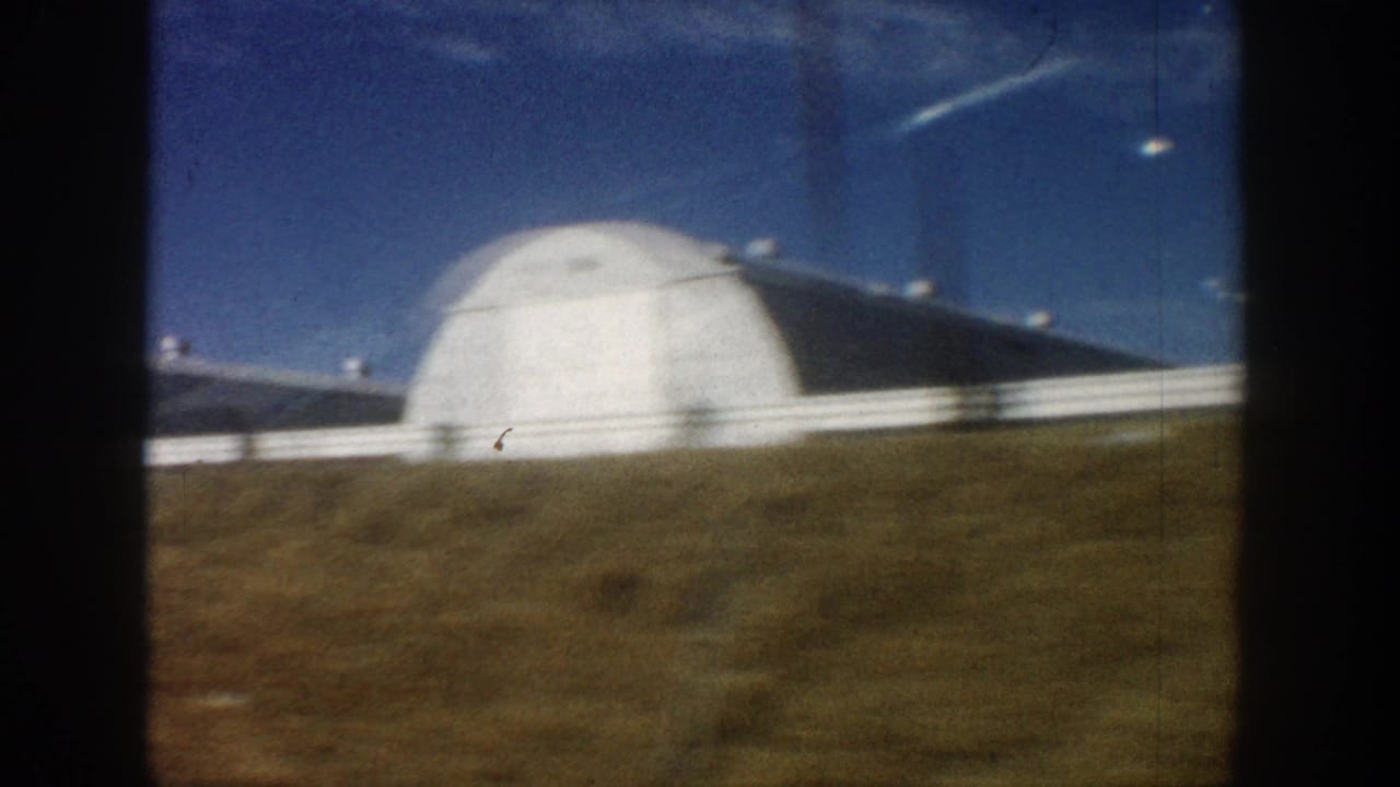 Large white dome structure, possibly a Quonset hut, filmed from a passing vehicle with dry scrubland and white fence rail visible; metadata records location as Colorado, 1961.