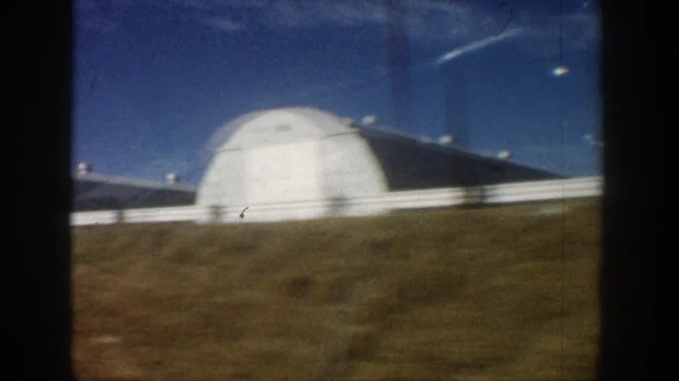 Large white dome structure, possibly a Quonset hut, filmed from a passing vehicle with dry scrubland and white fence rail visible; metadata records location as Colorado, 1961.