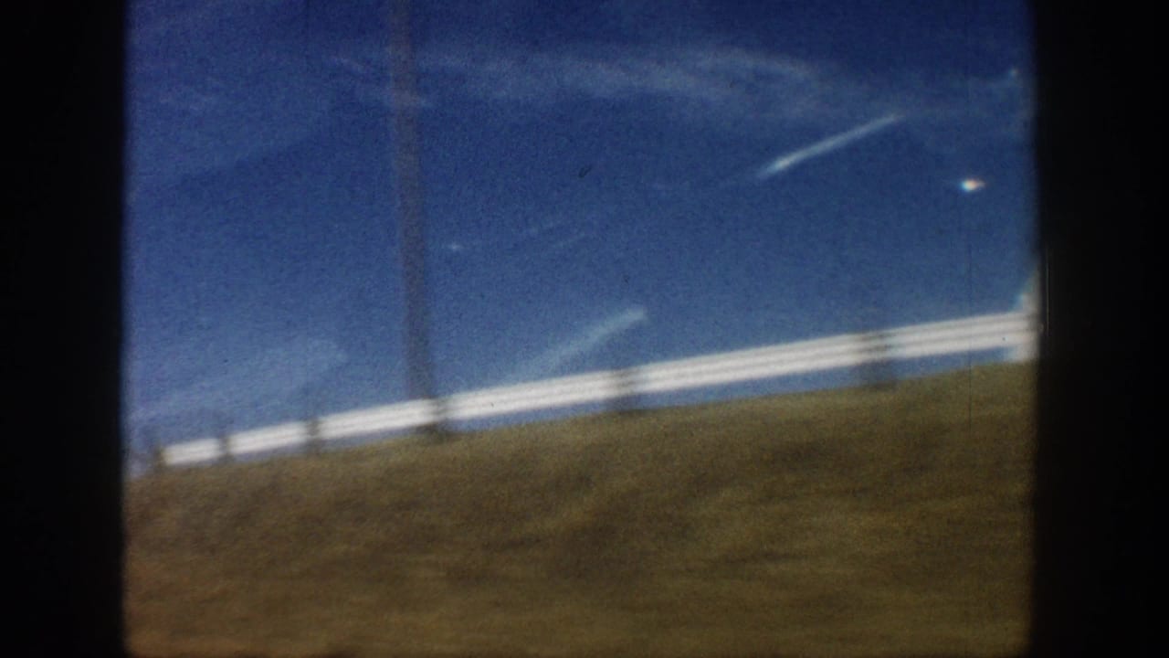 Empty flat scrubland with white painted fence rail under a broad blue sky, filmed from a moving vehicle in what metadata records as Colorado, 1961; dome structure has passed out of frame.
