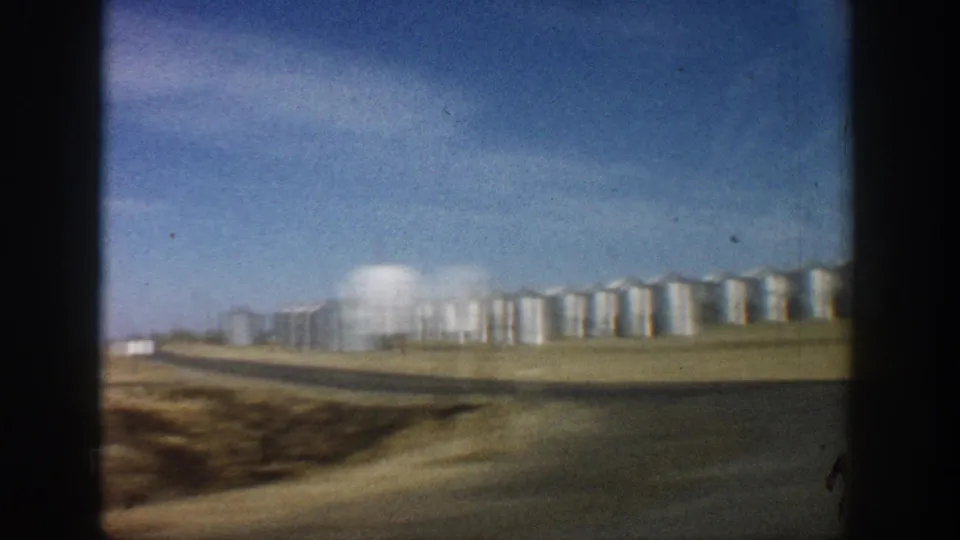 Long row of cylindrical metal grain silos alongside a white dome building on flat open terrain, filmed from a passing vehicle in what metadata records as Colorado, 1961.