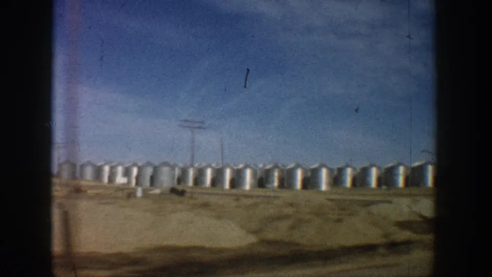 Extensive row of cylindrical metal grain silos stretching across flat landscape with utility poles, filmed from a moving vehicle circa 1961; slight film light-leak visible at right edge.