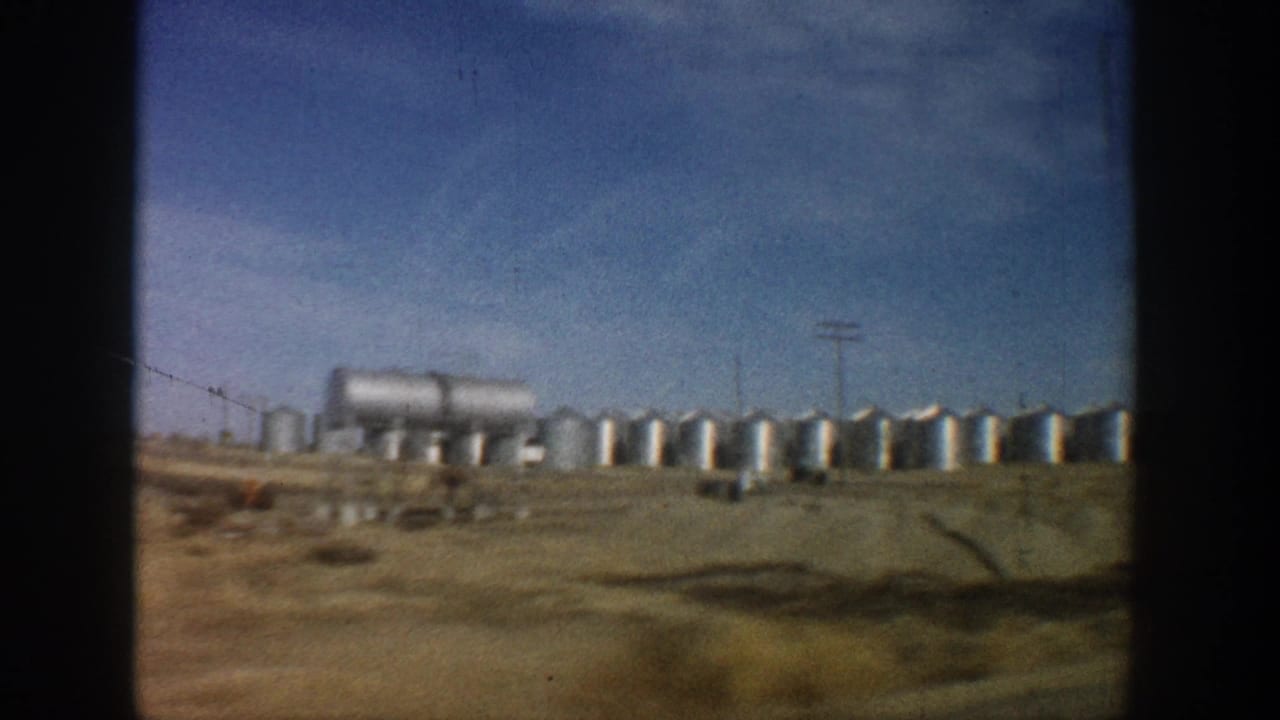 8mm color shot of industrial storage tanks and silos on a flat Colorado highway corridor plain under a clear blue sky, circa 1961.