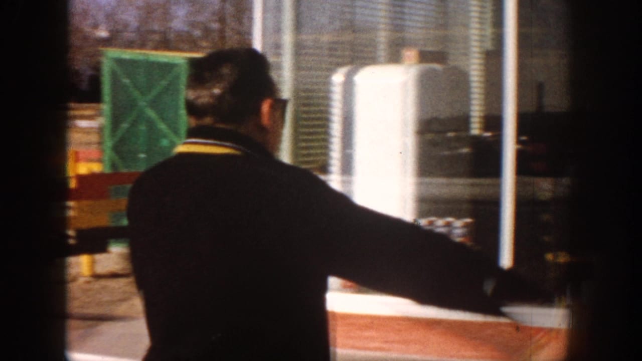 Man in dark jacket with yellow trim seen from behind at a store counter, white cylindrical storage tank and green crate visible in background.