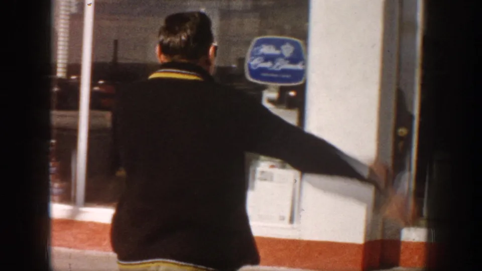 Man extending arms at a roadside storefront with a blue oval branded sign on the white facade, circa 1961 Colorado.