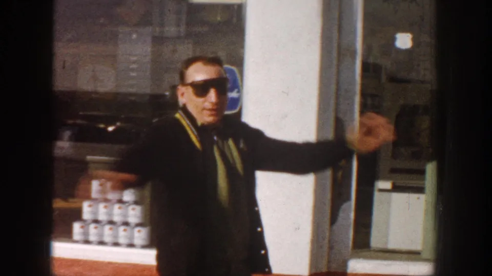 Man in dark-framed sunglasses and varsity-style jacket gestures toward camera outside a storefront, stacked cans and blue sign visible in window, circa 1961.