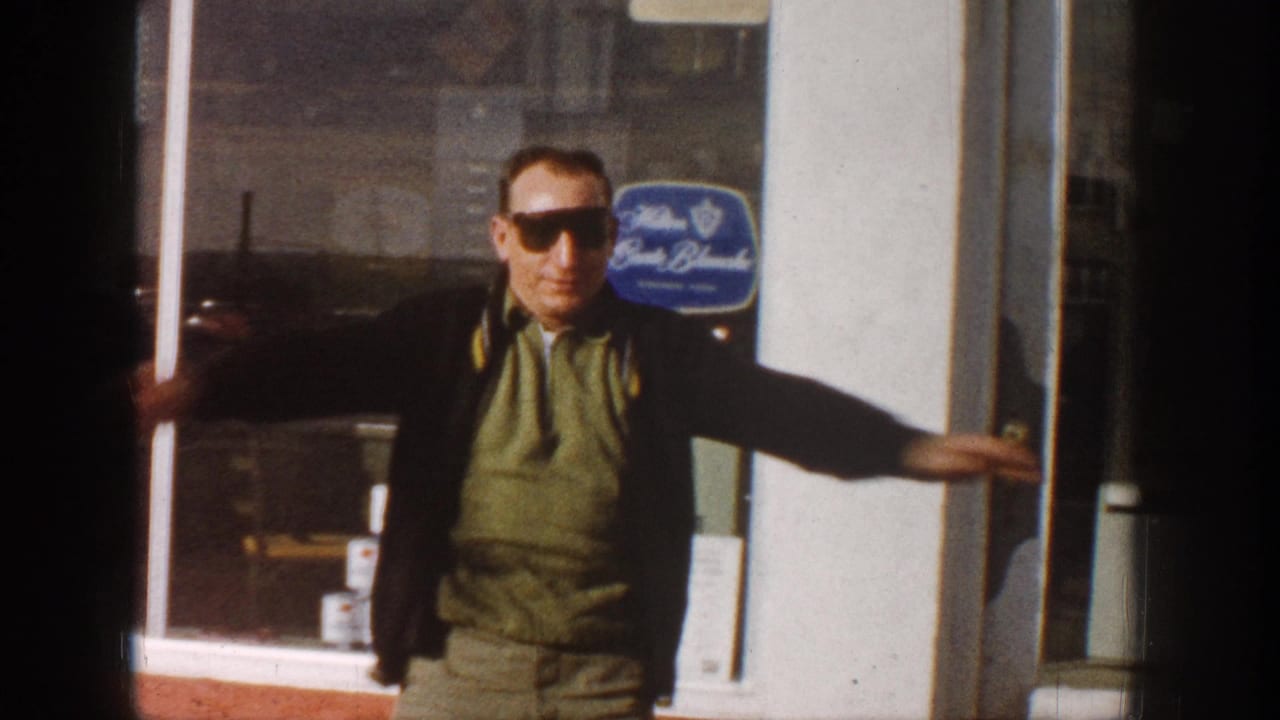 Man in sunglasses and olive-green vest stands at a commercial building entrance, hand on doorframe; blue oval sign mounted on exterior wall.