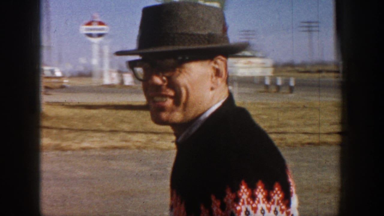 Man in dark fedora, thick-rimmed glasses, and red-and-white Nordic patterned sweater smiles outdoors; circular sign on tall pole and open flat terrain visible in background, circa 1961 Colorado.