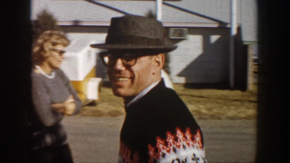 Man in dark fedora and red-and-white Nordic sweater faces camera with a smile; a woman with short light hair and sunglasses is partially visible at left, outdoors circa 1961.
