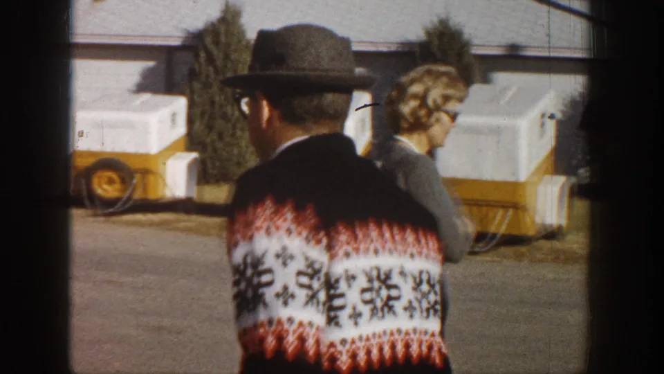 Rear view of man in dark fedora revealing the snowflake-motif back of his Nordic sweater; a woman stands to the right, with a yellow vehicle and white equipment cases visible in the background, circa 1961.