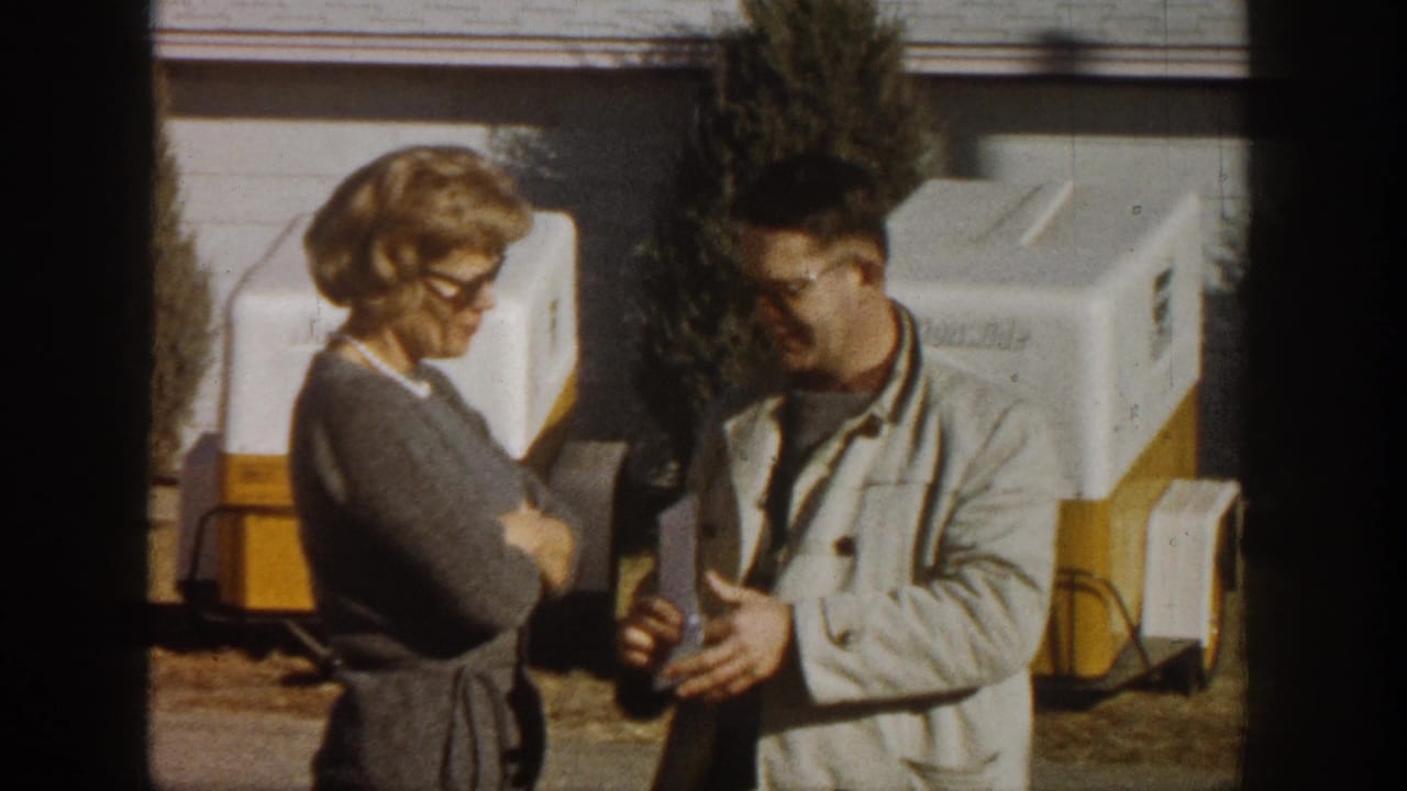 Woman in eyeglasses and gray sweater converses with man in light jacket holding papers; white and yellow trailer units behind them.