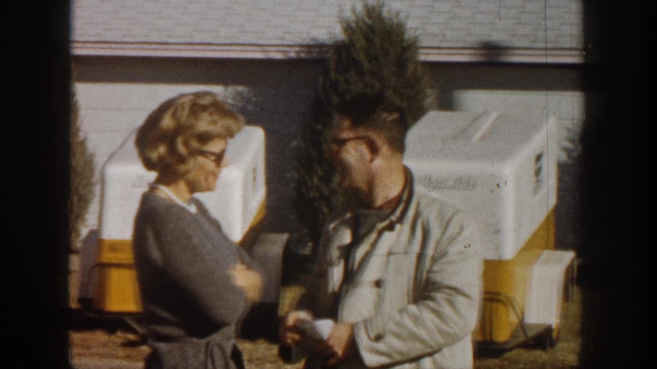 Woman turned in profile and man facing camera during outdoor conversation; portable white-and-yellow equipment units in background; Aspen Colorado 1961 8mm color footage.