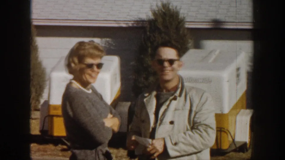 Man and woman both now wearing sunglasses smiling at camera in sunny outdoor setting; white-and-yellow equipment trailers behind them; Aspen Colorado 1961 8mm footage.