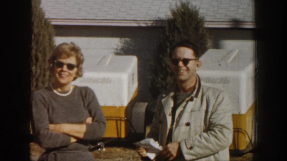 Close two-shot of smiling man and woman in sunglasses; papers visible in man's hand; white-and-yellow equipment trailers in background; 1961 8mm color film.