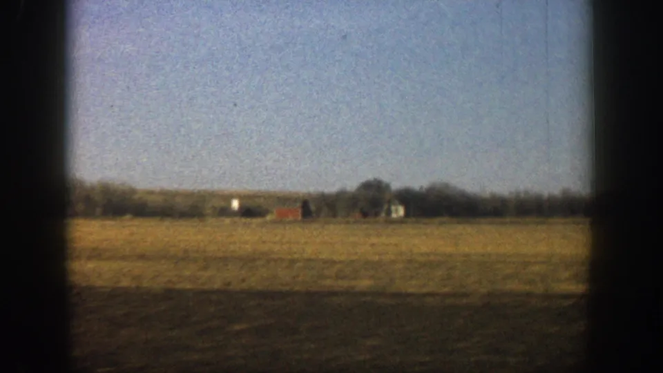 Wide shot of flat open plain with tan dry grass, a small red outbuilding, and additional farm structures on the far horizon; rural Colorado, circa 1961.