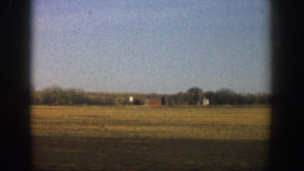 8mm home movie frame of open flat farmland with a distant red barn and farm structures at the horizon, shot through vehicle window, reportedly Colorado, circa 1961.
