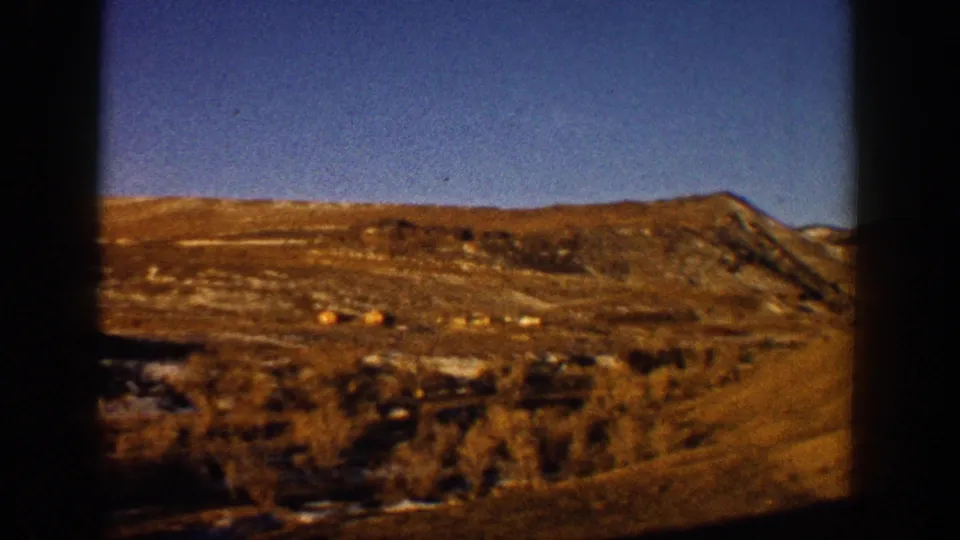 A steep eroded reddish hillside with what may be small structures or debris at its base, filmed through a car window, consistent with early 1960s 8mm Colorado footage.