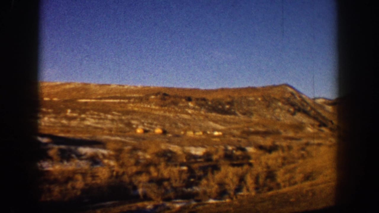 8mm home-movie frame from 1961 showing a snow-dusted Colorado hillside through a car window, golden scrub vegetation below a clear blue sky.