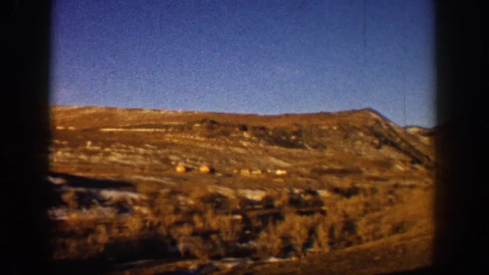 8mm home-movie frame from 1961 showing a snow-dusted Colorado hillside through a car window, golden scrub vegetation below a clear blue sky.