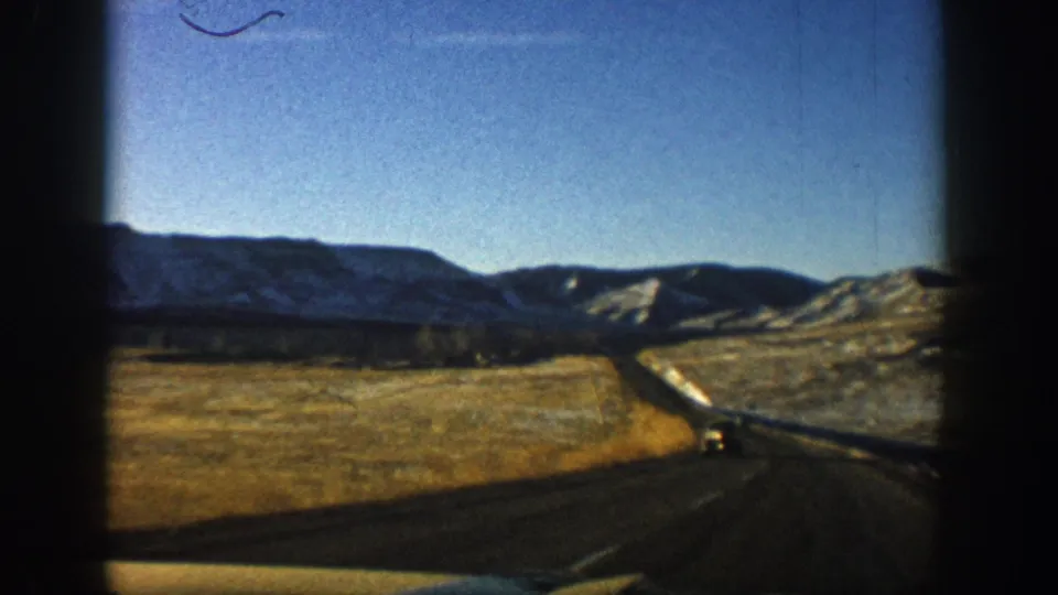 8mm home-movie frame from 1961 showing a two-lane Colorado highway through a mountain valley, snow-dusted ridges in the background and a distant vehicle on the road.
