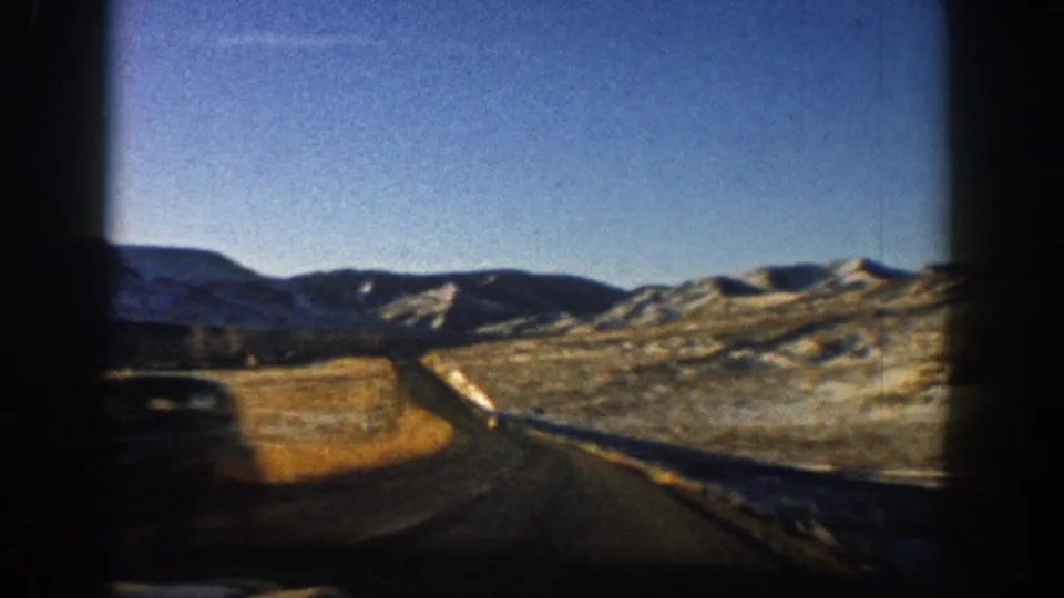 8mm home-movie frame from 1961 showing a two-lane Colorado highway curving through a valley, snow-dusted rolling hills and clear blue sky visible.