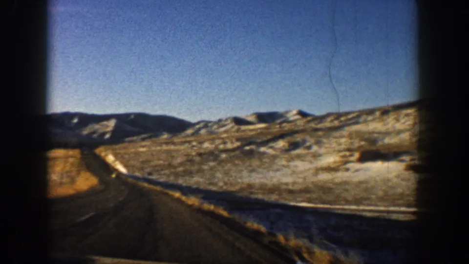 8mm home-movie frame from 1961 showing a Colorado mountain highway and snow-dusted ridges across an open valley; a thin film-scratch artifact is visible in the sky.