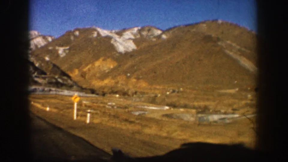 8mm film frame from moving car looking ahead along a straight mountain highway with snow-capped peaks, Colorado Rockies area, circa 1961.