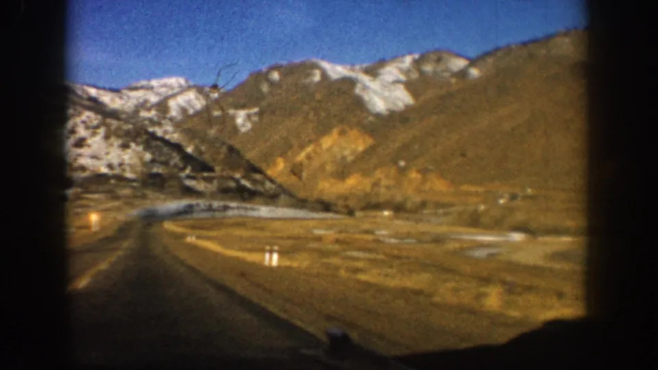 8mm home movie frame looking down a rural Colorado mountain highway with bare branches at right and snow-capped peaks in background, circa 1961.